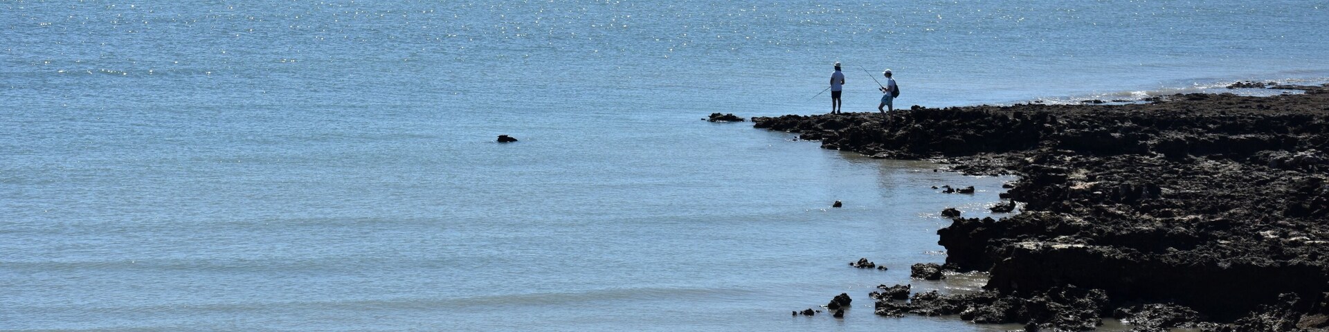 Una costa rocosa con dos personas pescando, mar tranquilo y cielo despejado.