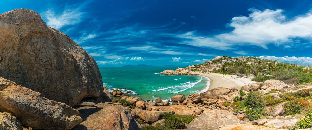 A rocky coastline with a large boulder, turquoise sea, and a sandy beach.
