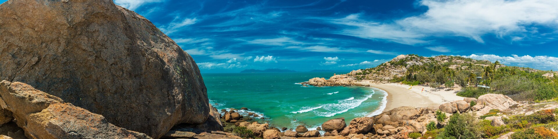 A rocky coastline with a large boulder, turquoise sea, and a sandy beach.