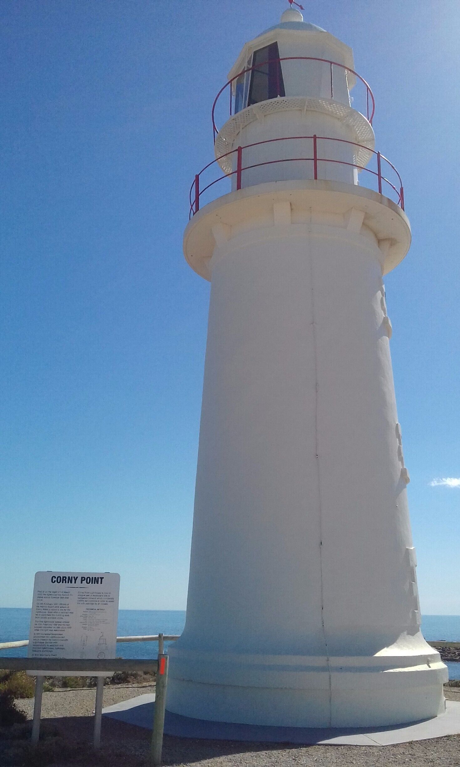 A white lighthouse with a sign reading "Corny Point" in front of a clear blue sky.