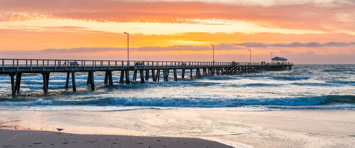 A pier extending into the ocean with waves crashing against it, a clear sky, and a sunset.