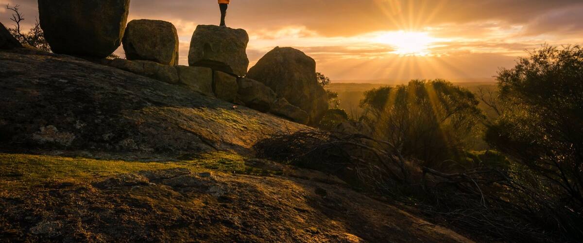 A person standing on a rock formation with the sun setting in the background.