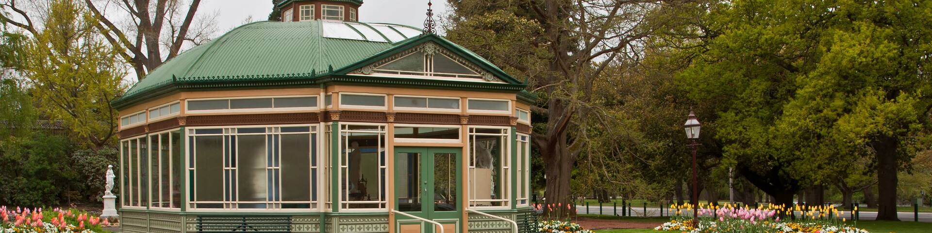 A gazebo with a green roof, surrounded by a garden with various colorful flowers and trees.