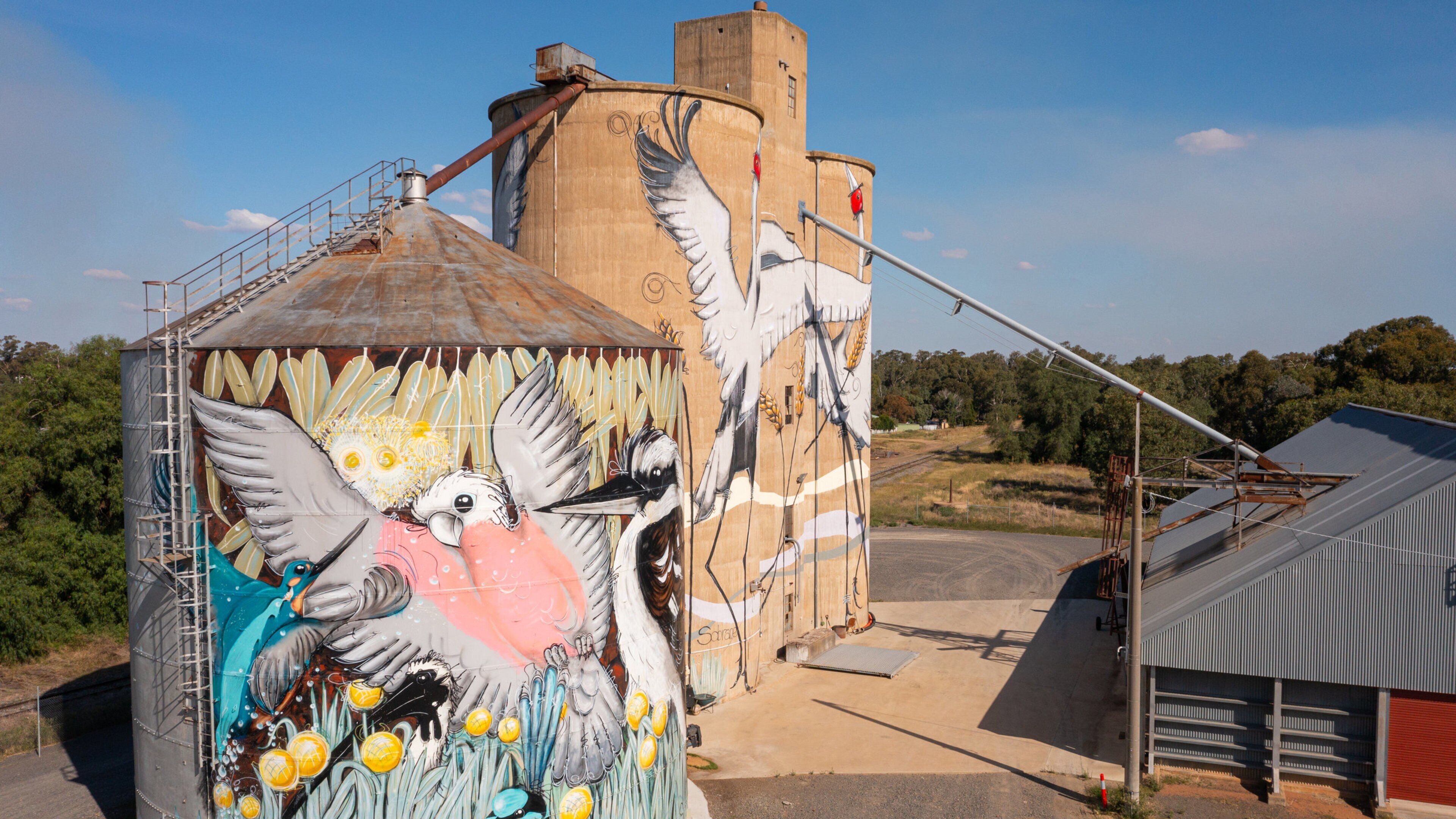 A large grain silo with a mural of birds and a silo with a metal roof.
