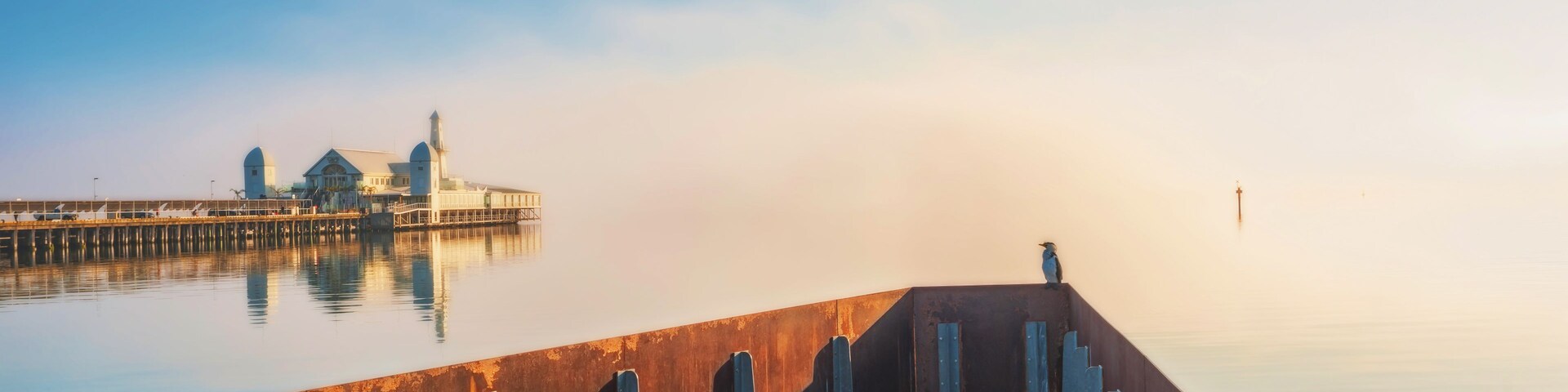 A wooden pier extending into calm water with a foggy backdrop and a distant building.