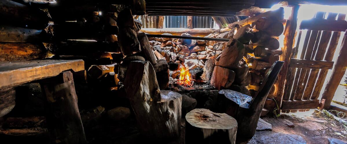 A rustic outdoor cooking area with a fire, wooden logs, and a metal pot.