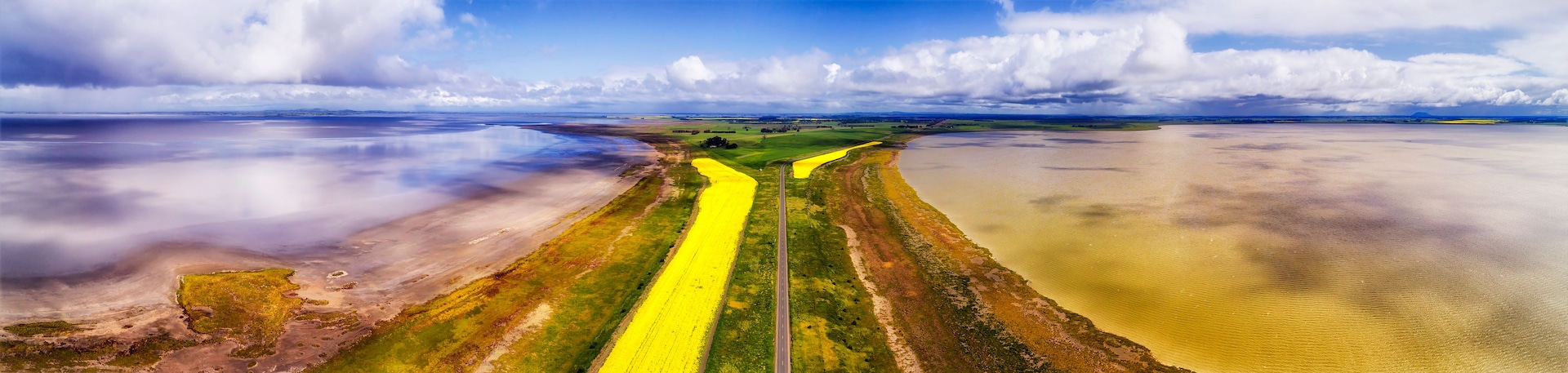 Un vaste paysage dominé par un champ jaune éclatant, une étendue d’eau et un ciel nuageux.