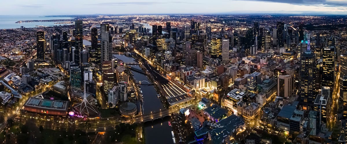 A cityscape at dusk with illuminated buildings, a river, and a distant coastline.