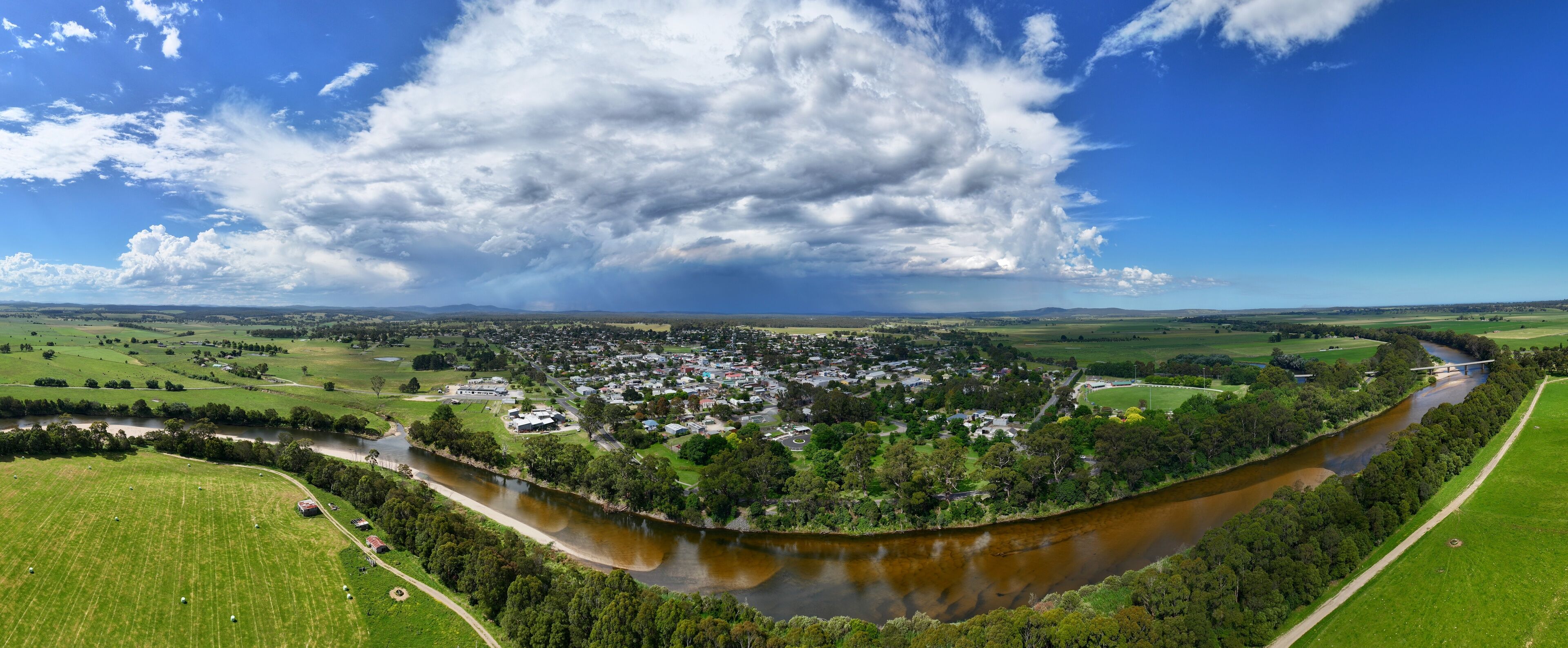 A river winding through a town surrounded by green fields and a large cloud in the sky.
