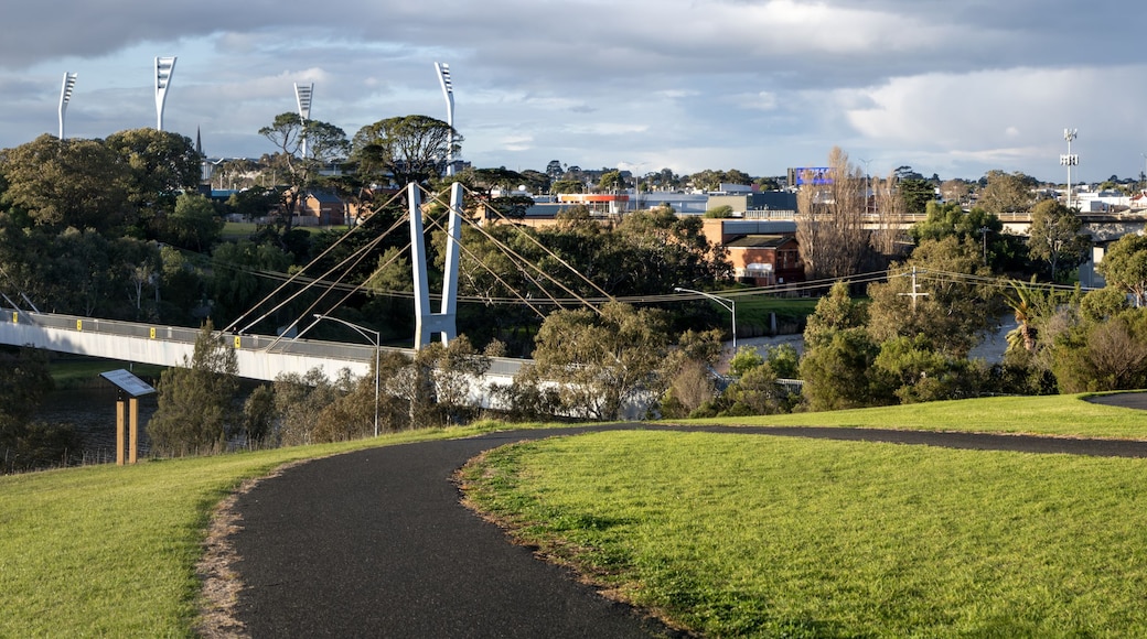 Walking trail across grassy hills towards a pedestrian bridge over the Barwon River in Geelong, Victoria, Australia. Panoramic view of green parkland and regional city skyline.
