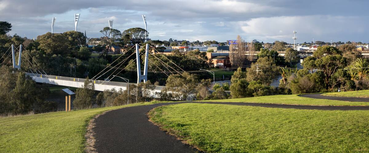 Walking trail across grassy hills towards a pedestrian bridge over the Barwon River in Geelong, Victoria, Australia. Panoramic view of green parkland and regional city skyline.