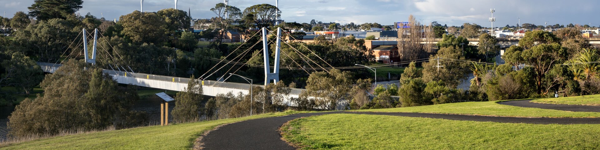 A pedestrian bridge over a river, surrounded by greenery and urban buildings.