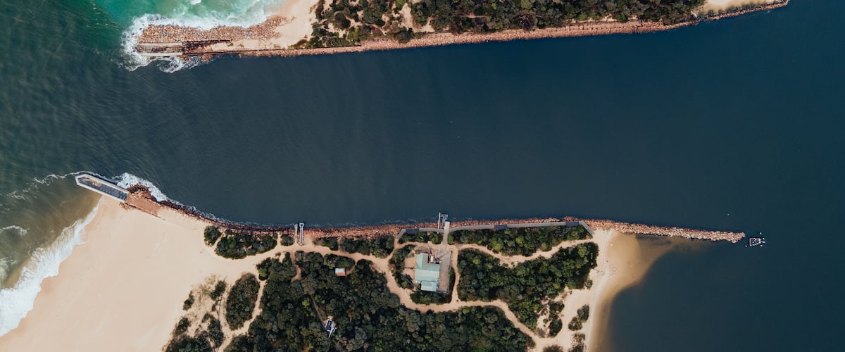 An aerial view of a coastal area with a beach, a forested peninsula, and a long pier extending into the water.