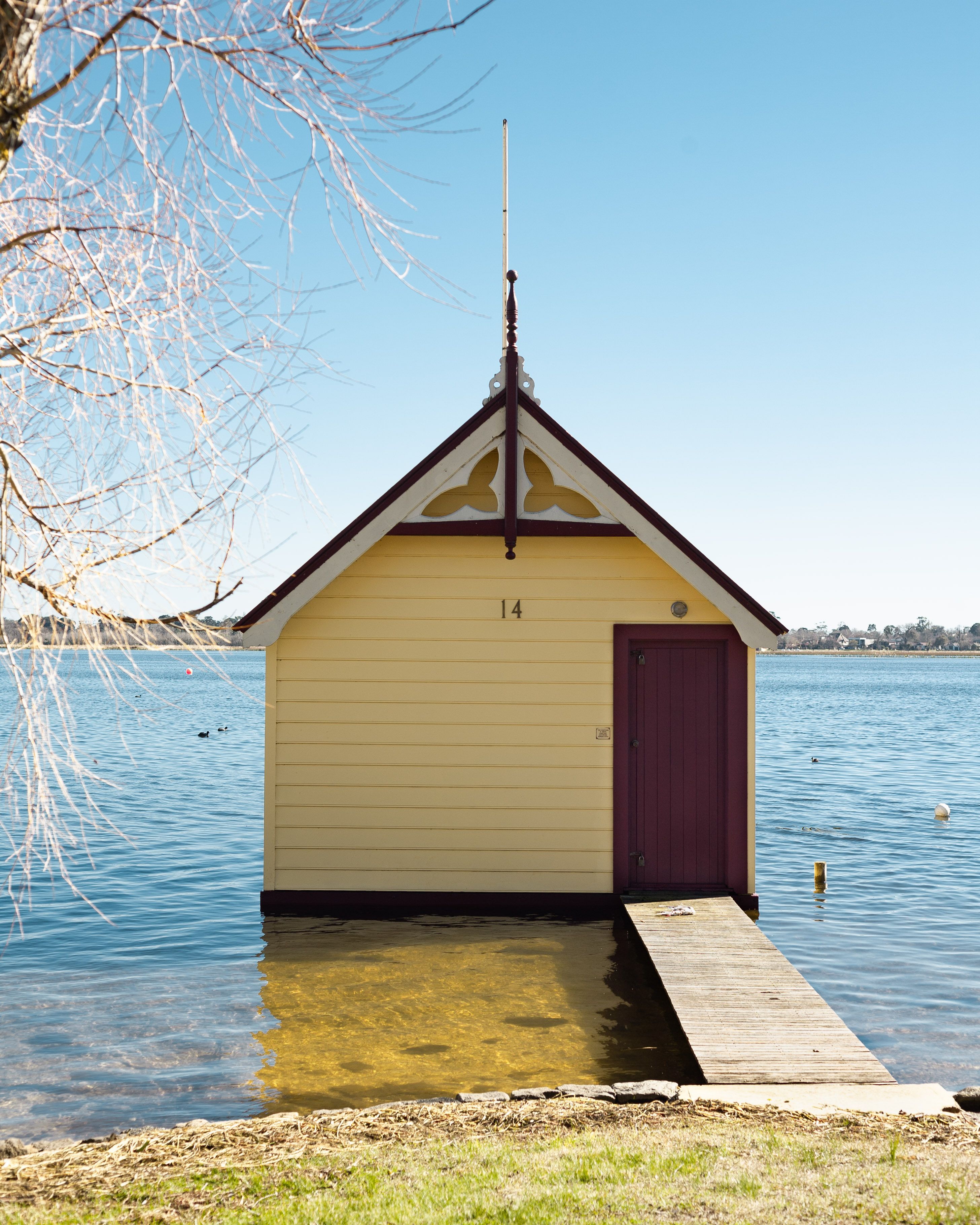 Uma pequena casa de barcos amarela com uma porta roxa, número 14, em um píer que se estende sobre um lago calmo.