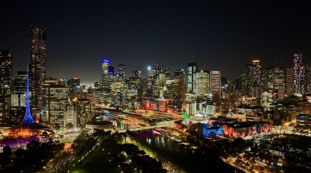 A nighttime cityscape with illuminated skyscrapers, a prominent tower with blue lights, and a river with colorful lights reflecting on the water.