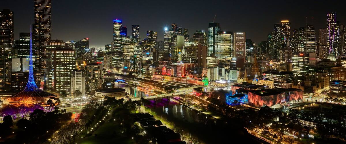 A nighttime cityscape with illuminated skyscrapers, a prominent tower with blue lights, and a river with colorful lights reflecting on the water.