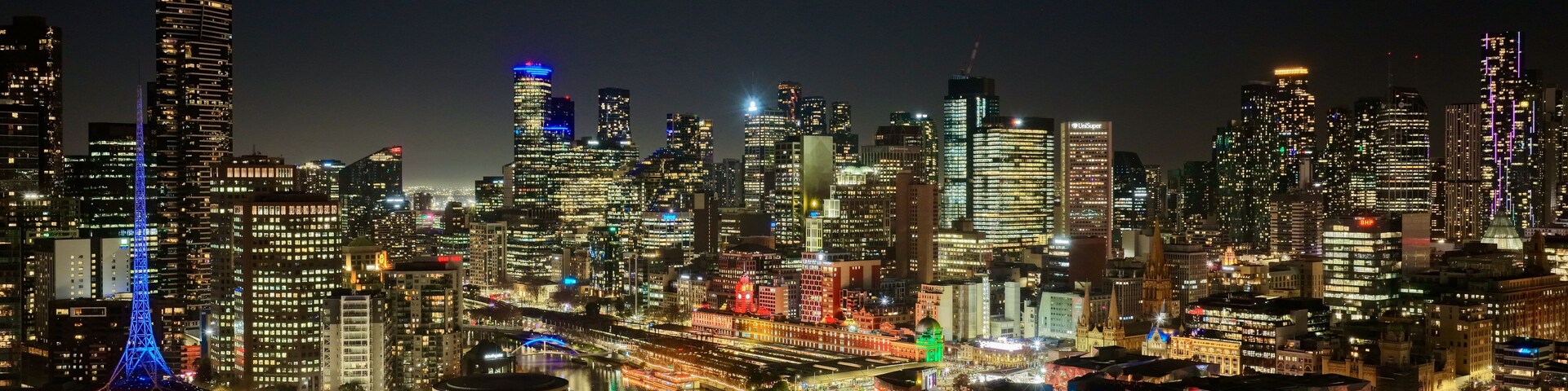 Paisagem urbana noturna com arranha-céus iluminados, uma torre de destaque com luzes azuis e um rio com reflexos coloridos na água.