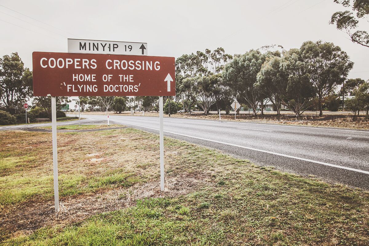 A road sign indicating 'Coopers Crossing' and 'Home of the Flying Doctors' with an arrow pointing straight ahead.