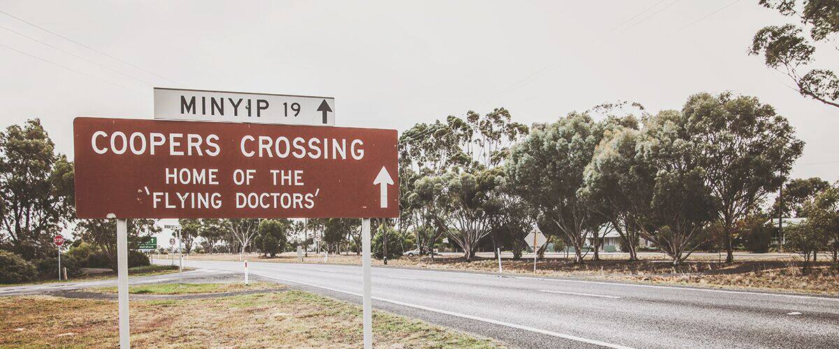 A road sign indicating 'Coopers Crossing' and 'Home of the Flying Doctors' with an arrow pointing straight ahead.