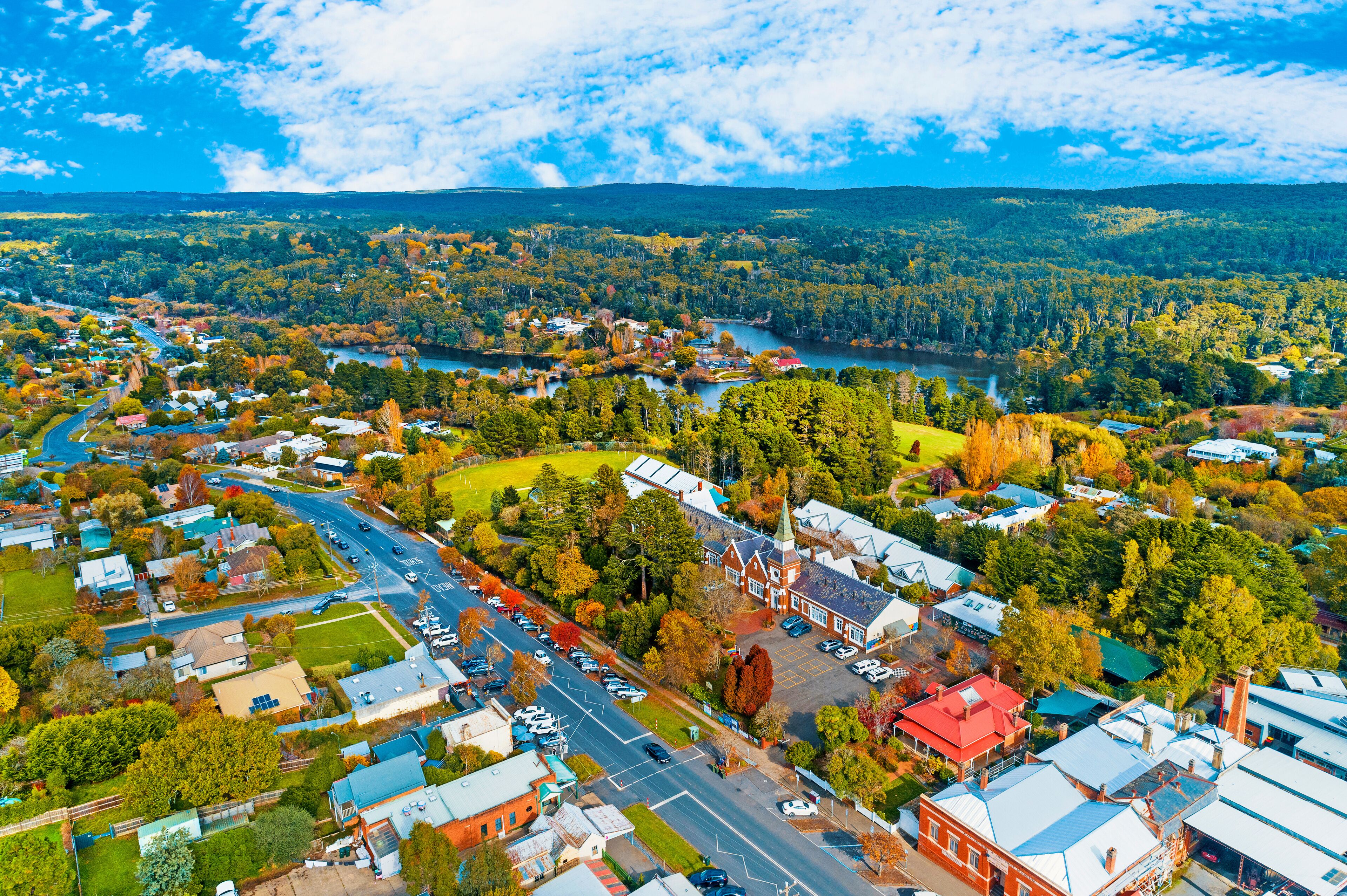 An aerial view of a town with a river, green hills, and autumn-colored trees.