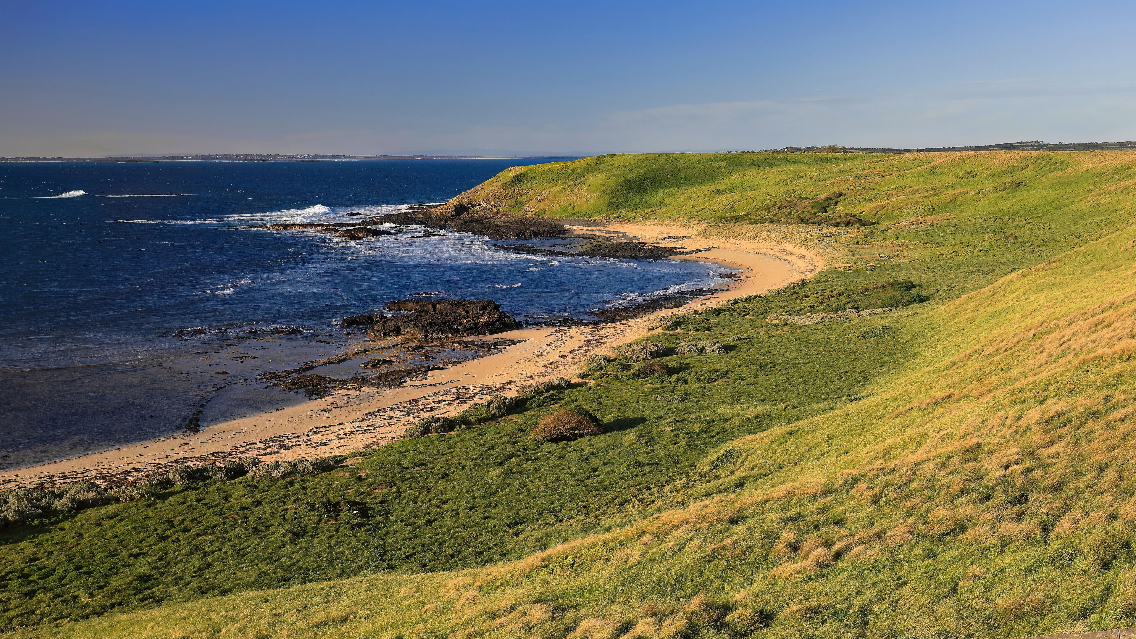 A coastal landscape with a sandy beach, rocky shoreline, and green hills.