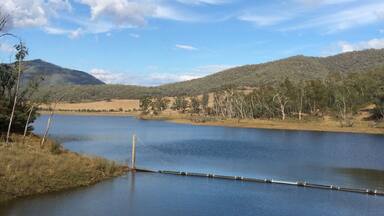 A calm lake with a wooden dock, surrounded by hills and sparse trees under a partly cloudy sky.
