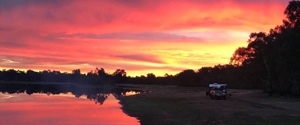 A sunset over a lake with a reflection, a dirt path, and a vehicle parked on the right.