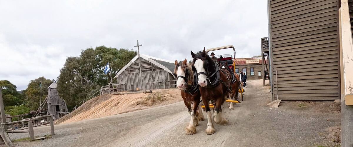 A dirt road with two horses pulling a cart, a wooden building, and a church with a cross.