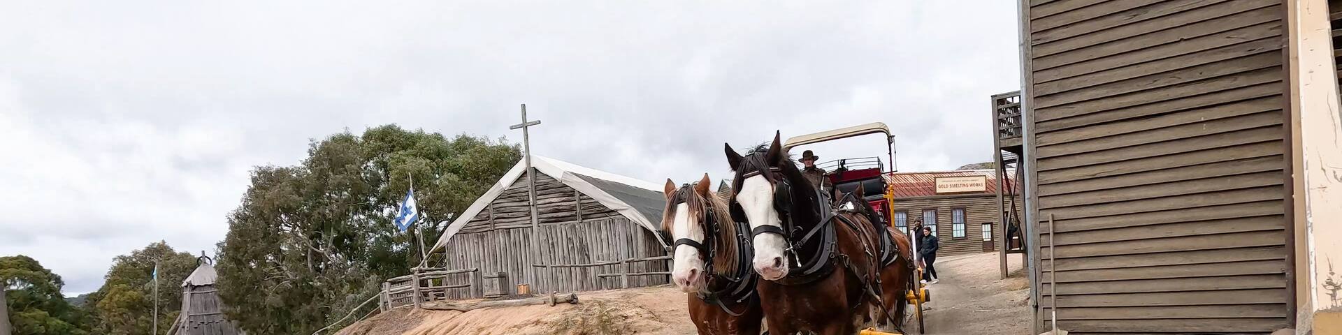 A dirt road with two horses pulling a cart, a wooden building, and a church with a cross.