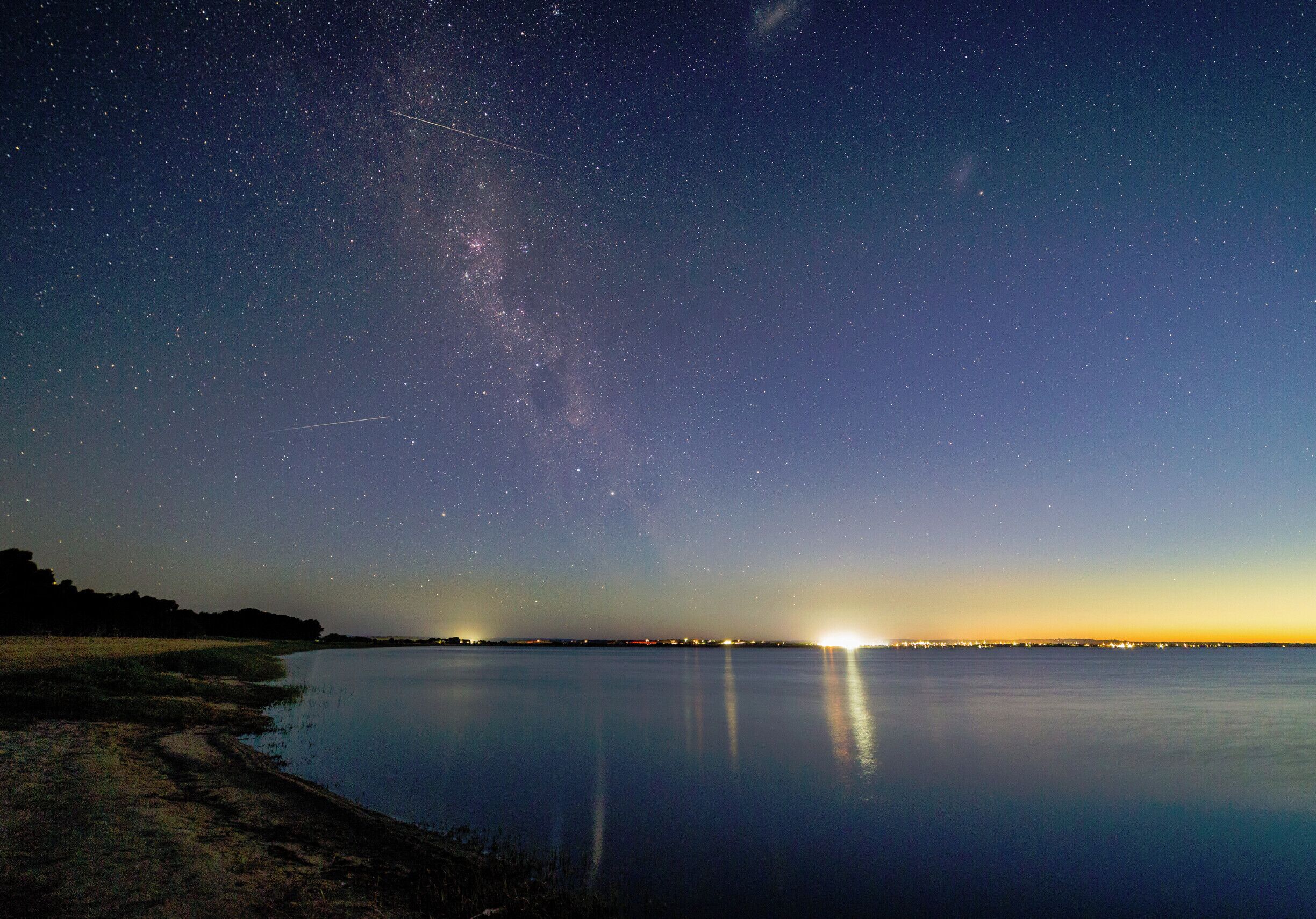 A starry night sky over a calm body of water with a visible Milky Way and city lights in the distance.