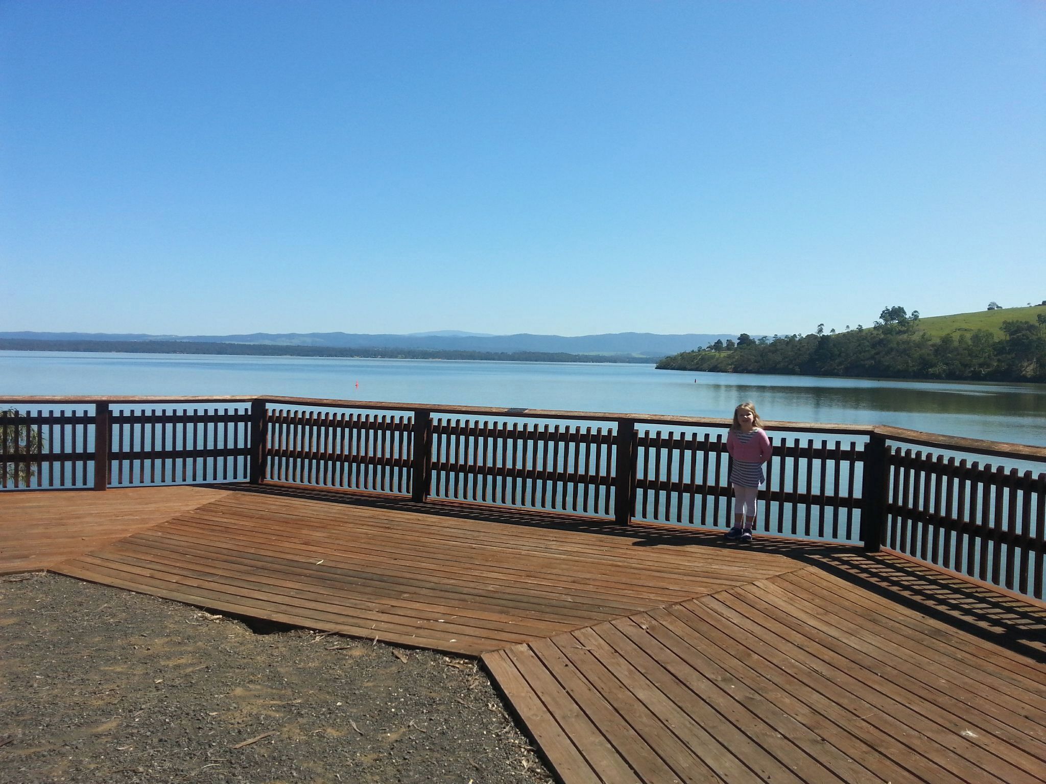 A wooden pier with a railing overlooking a calm lake, surrounded by hills and trees.