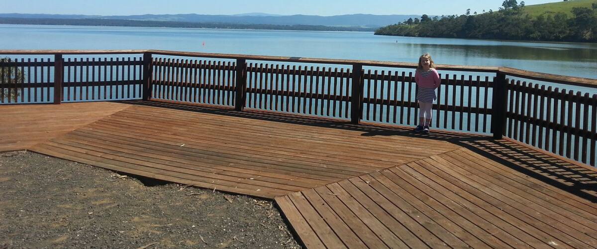A wooden pier with a railing overlooking a calm lake, surrounded by hills and trees.