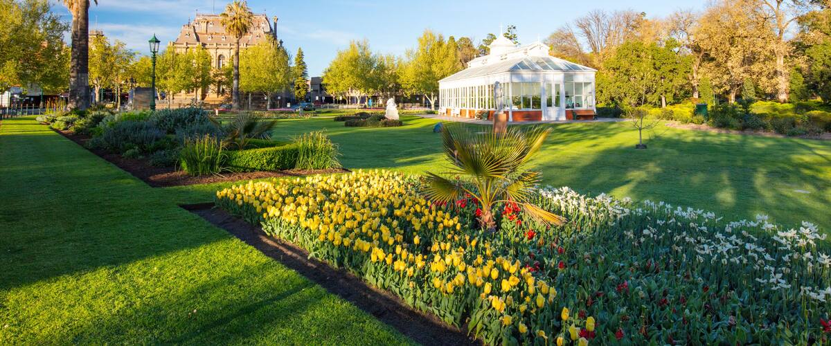 A well-maintained garden with a variety of flowers, a large white building, and a historic building in the background.