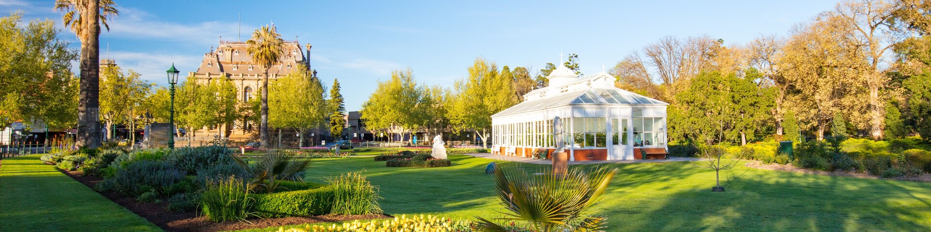 Un jardín bien cuidado con diversas flores, un edificio blanco grande y un edificio histórico al fondo.