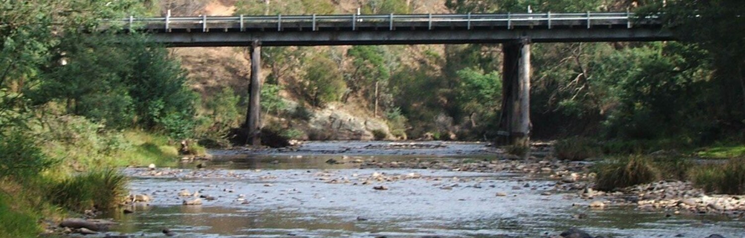 Un puente sobre un río con riberas rocosas y una ladera boscosa al fondo.