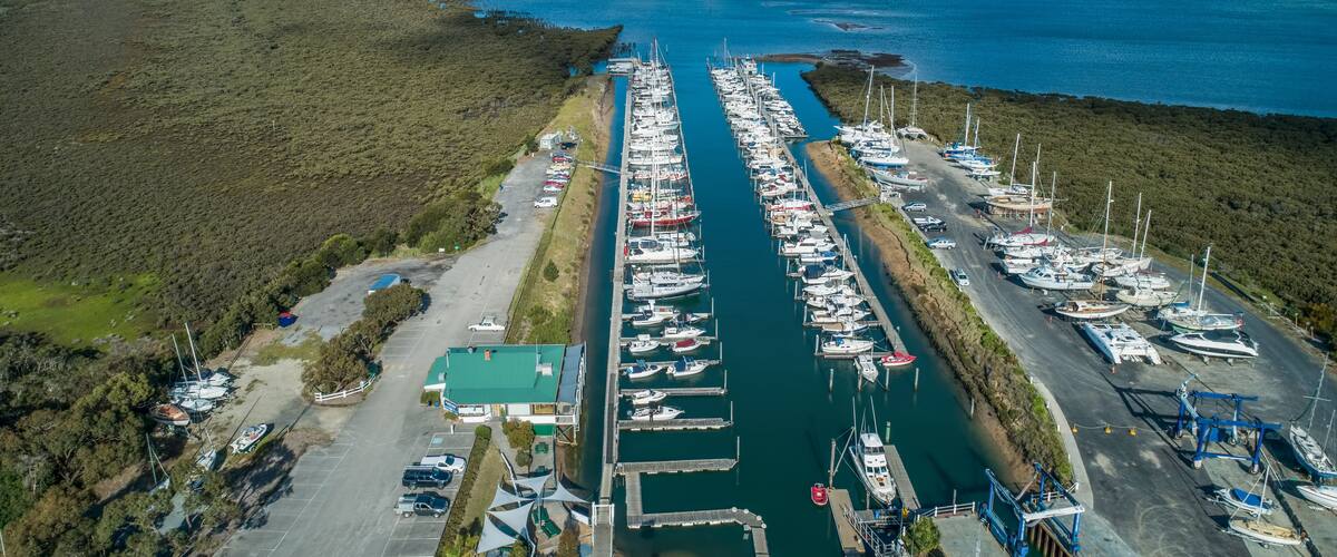 Vista aérea de un puerto deportivo con numerosas embarcaciones amarradas a lo largo de un canal estrecho, rodeado de vegetación densa y un cielo despejado.