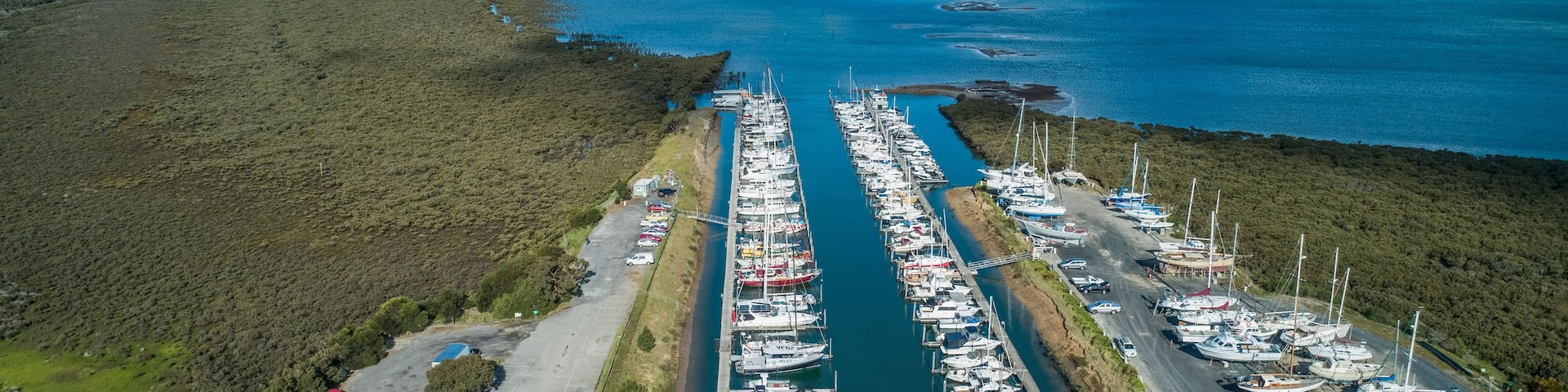Vista aérea de un puerto deportivo con numerosas embarcaciones amarradas a lo largo de un canal estrecho, rodeado de vegetación densa y un cielo despejado.
