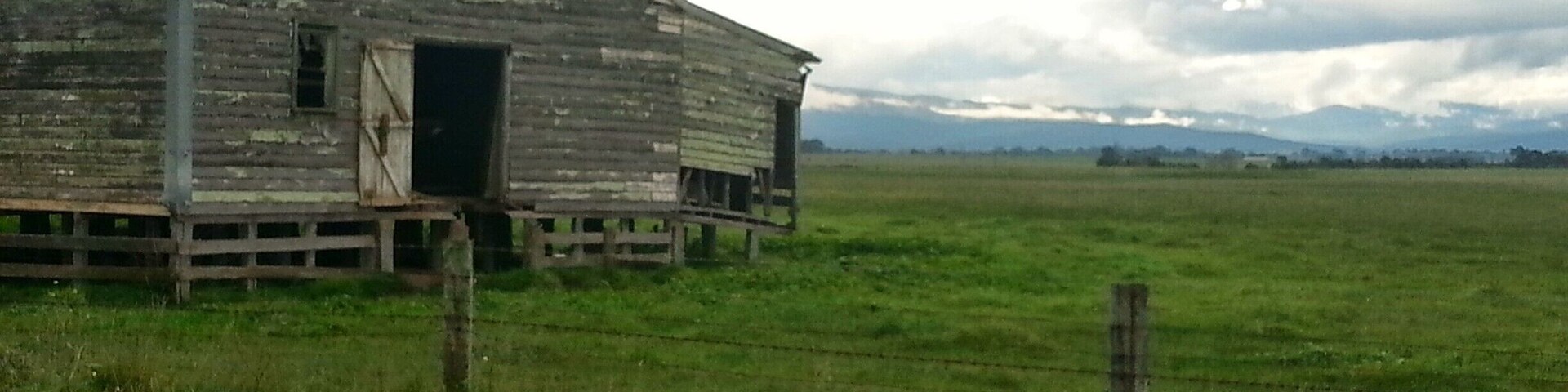 An old wooden barn with a metal roof, surrounded by a grassy field and a barbed wire fence.