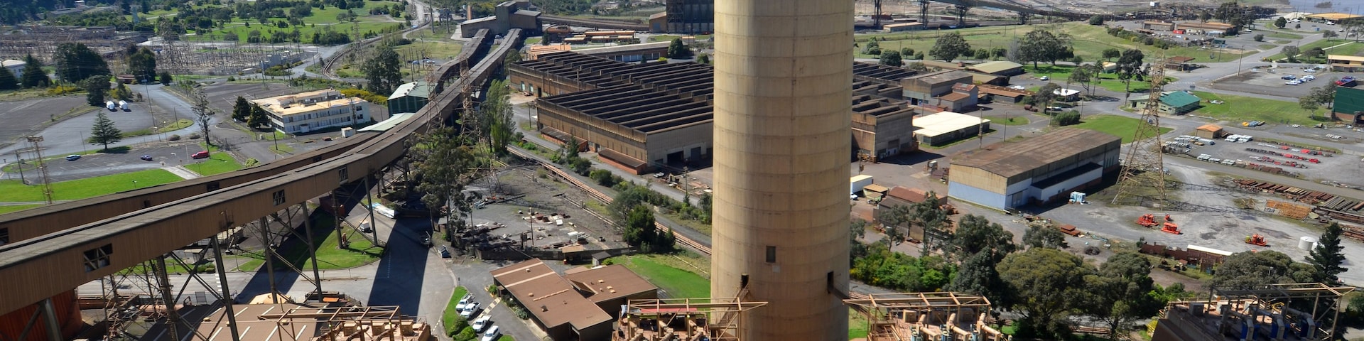 Um grande complexo industrial com uma torre de resfriamento imponente, trilhos de trem e campos verdes ao redor.
