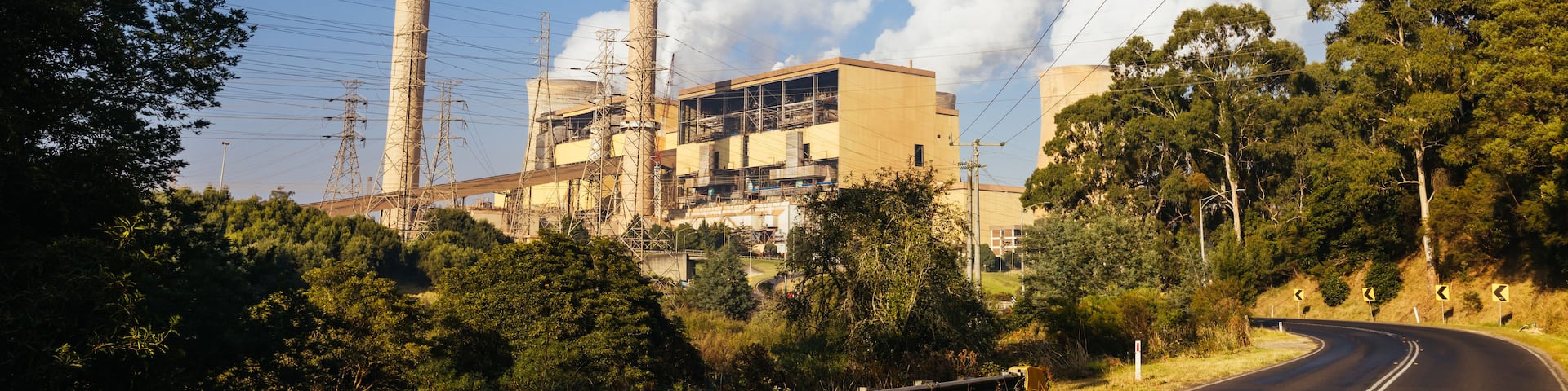 A road curving through a landscape with a large industrial facility with multiple chimneys and power lines overhead.