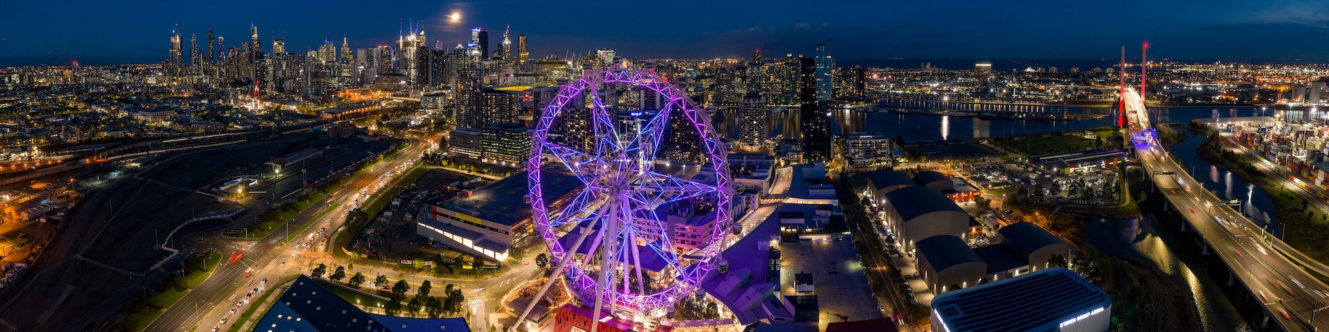 Paisagem urbana noturna com uma roda-gigante e uma ponte em destaque.