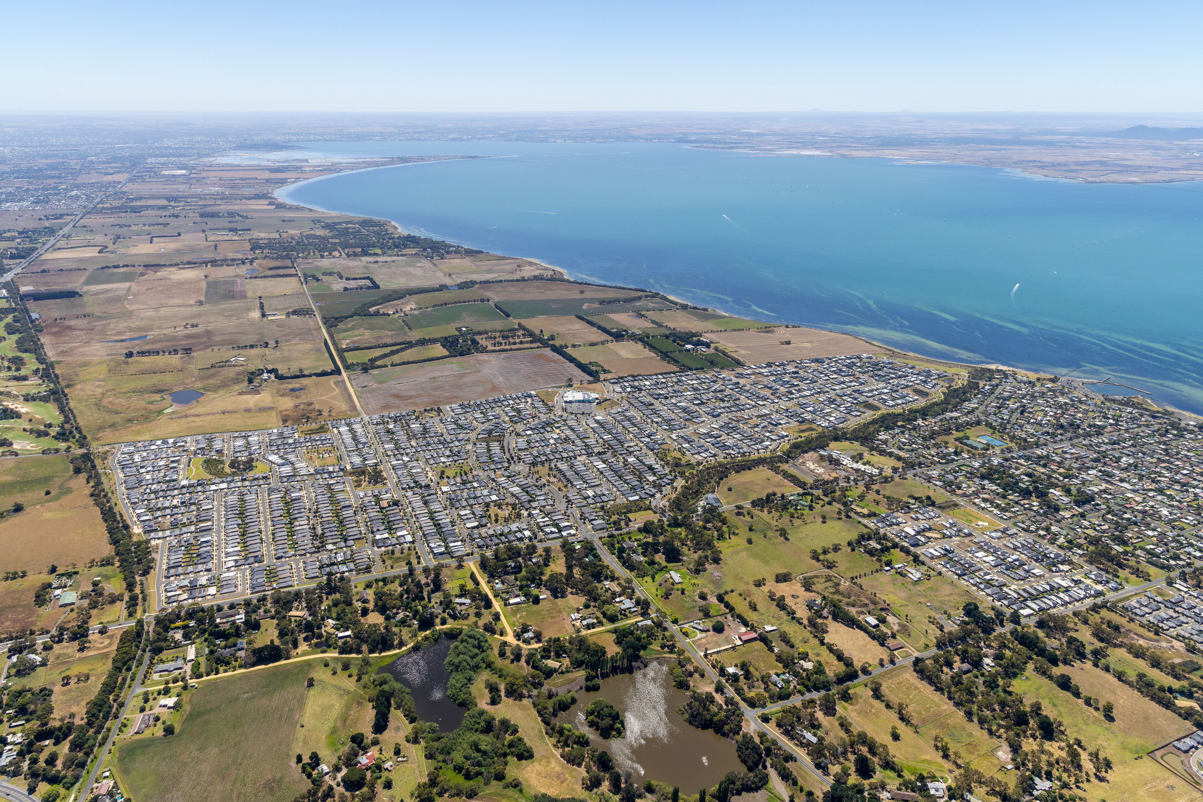 An aerial view of a coastal town with residential areas, a large body of water, and surrounding farmland.