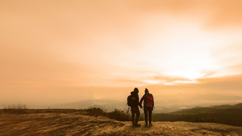 Two hikers on a rocky outcrop overlooking a valley with a sunset in the background.
