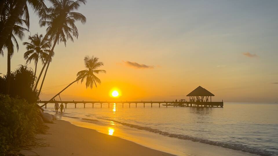 A beach at sunset with palm trees, a pier, and a small hut.
