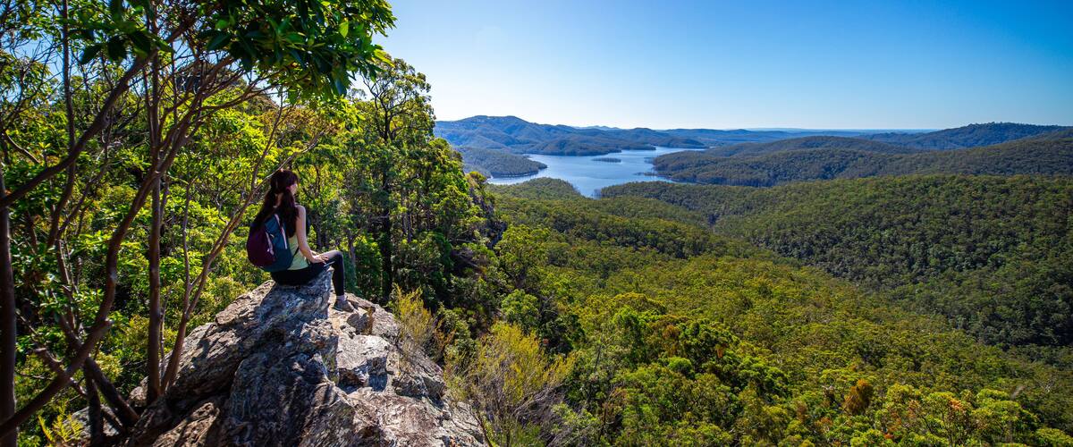 A person sitting on a rock overlooking a valley with a lake and hills.