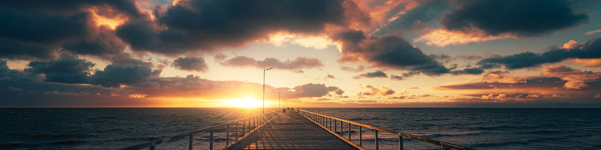 Um píer de madeira que se estende até o oceano, com o pôr do sol ao fundo.