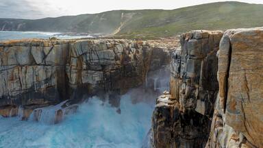 A coastal cliff with a waterfall, rugged terrain, and a distant view of the ocean.
