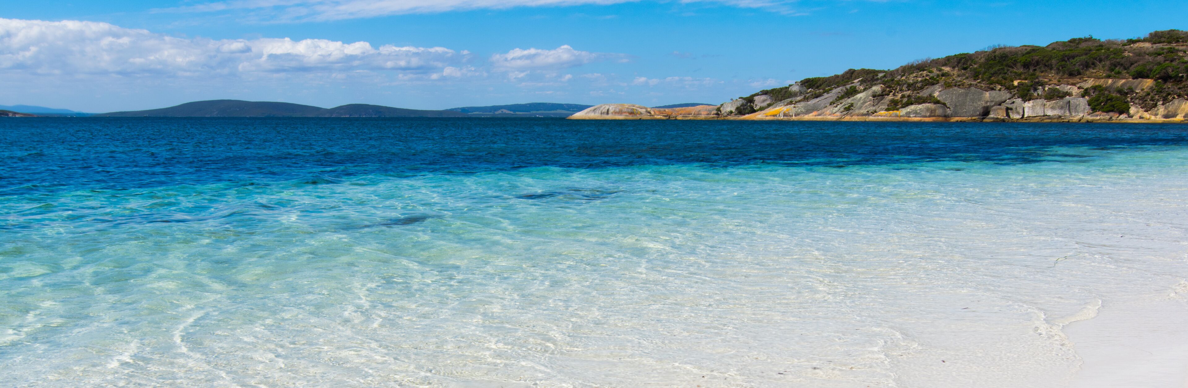 A beach with clear turquoise water and white sand, surrounded by rocky hills and sparse vegetation.
