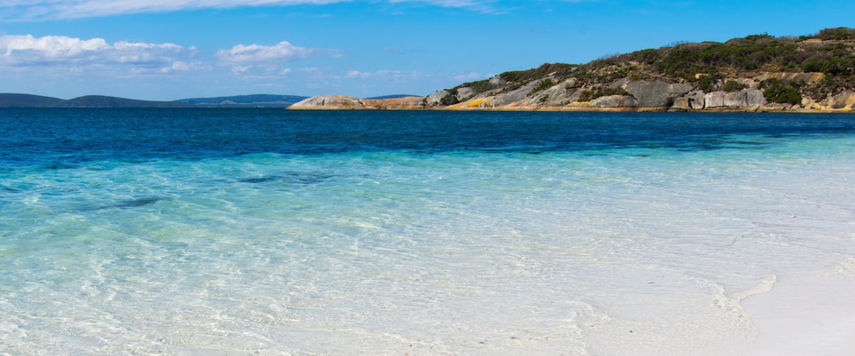 A beach with clear turquoise water and white sand, surrounded by rocky hills and sparse vegetation.