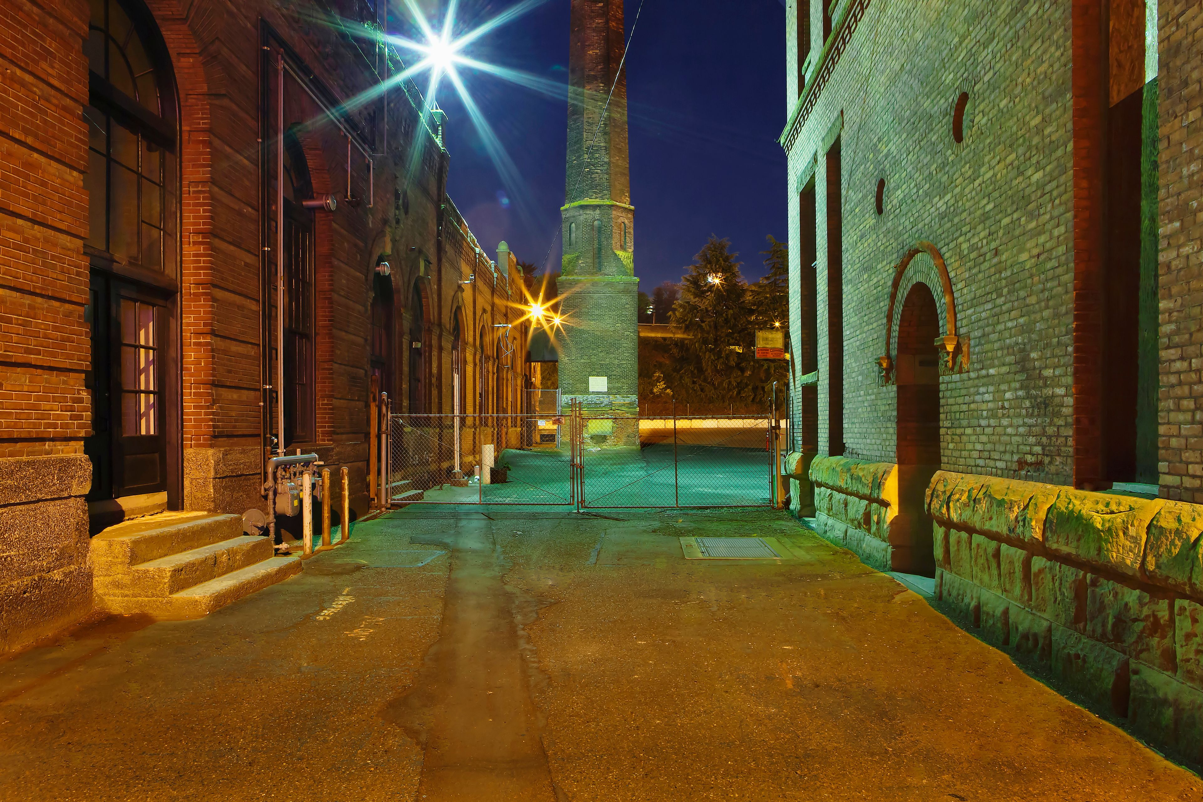 A nighttime street scene with brick buildings, a metal gate, and a tower in the background.